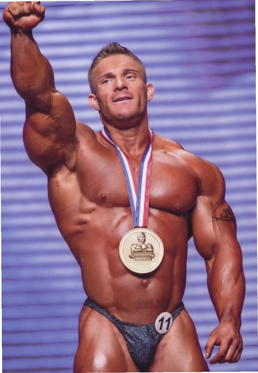 James Flex Lewis với huy chương vàng trên cổ, đứng trên sân khấu với cánh tay giơ cao