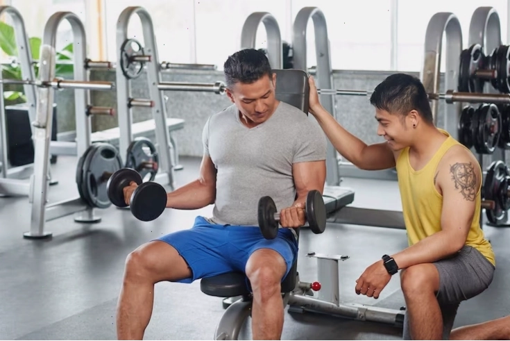 Vận động viên thể hình ngồi trên ghế tập tạ thực hiện nâng tạ đòn có kiểm soát bởi huấn luyện viên cá nhân